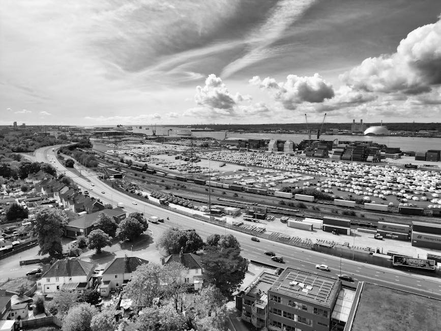A black and white aerial photograph showing an industrial port area near West Ham, with a large parking lot filled with numerous white vehicles, including trucks and containers, alongside railway tracks. In the foreground, a residential area with houses, trees, and streets is visible, with some moving equipment such as trolleys or small vehicles near the buildings. The sky above features clouds with streaks and puffs, and the scene includes part of a road running adjacent to the residential neighborhood, indicating the connection between home relocation activities and the logistics of furniture transport and packing in the West Ham E6 area. This image exemplifies the logistics involved in house removals, with a focus on the transportation and movement of furniture and boxes, as managed by services like Man and Van West Ham, specializing in efficient house removals near West Ham station.