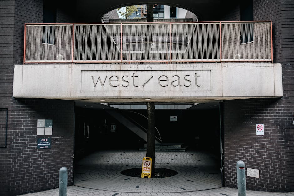 The image shows the entrance to a residential or commercial building in West Ham, E6, with a modern black brick façade and a concrete balcony area featuring a metal railing painted in a reddish-brown color. Beneath the balcony, there is a wide open archway leading into a parking or underpass area, with a concrete pillar situated directly in front of the entrance. Attached to the concrete section of the building is a sign displaying the words 'west / east' engraved into the concrete surface, indicating directional or location information. On either side of the entrance, there are small street signs and bollards protecting the area. Inside the archway, a staircase is partially visible, and the premises are illuminated with natural daylight, highlighting potential pathways for furniture or boxes related to home relocation or moving services. The scene depicts a typical environment for residential removals, with an emphasis on entry access for vehicle loading and unloading, supported by the presence of the potential moving logistics of a company like Man and Van West Ham.