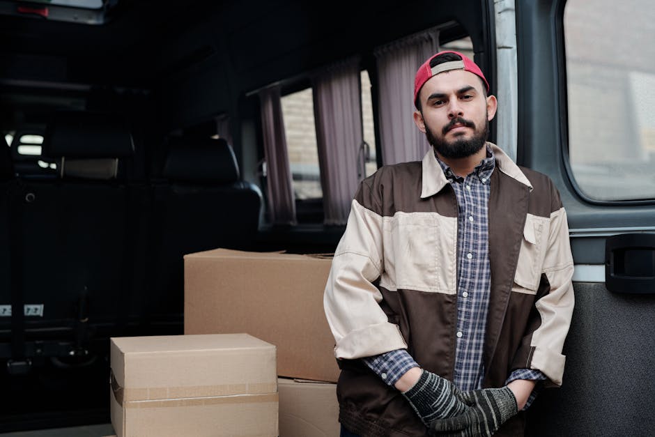 A young man with a serious expression is sitting on the pavement in front of the open rear of a dark van, which is parked outside a residential building. Inside the van, several cardboard moving boxes