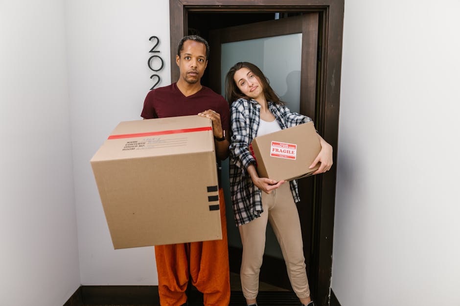 A cardboard box with its flaps open and partially sealed is positioned upright on a bed in a room with wooden paneling on the wall and a white curtain above a window. To the left of the box, there is