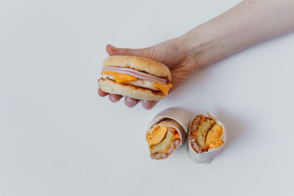 A person's hand is holding a breakfast sandwich featuring a soft bread roll filled with a slice of ham, a fried egg, and melted cheddar cheese, positioned above a white surface. Below, there are two p
