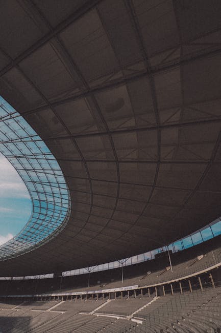 The interior of a large stadium or arena with a curved, modern roof structure featuring multiple panels and a circular opening revealing a blue sky with some clouds. The seating area below consists of