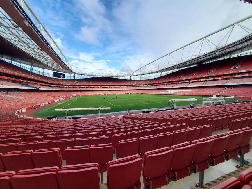 The interior of a large stadium or arena with a curved, modern roof structure featuring multiple panels and a circular opening revealing a blue sky with some clouds. The seating area below consists of