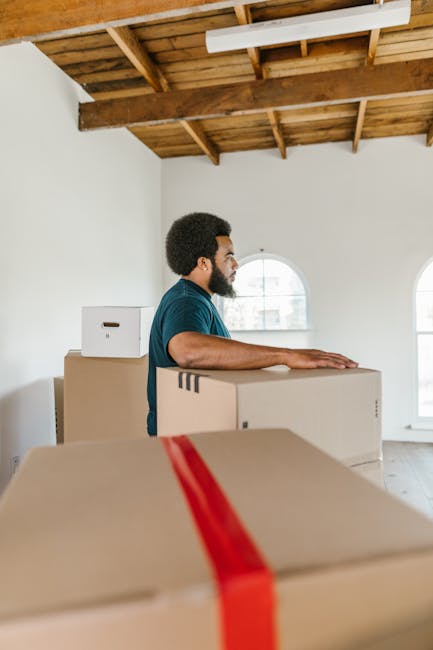 A man with curly dark hair, wearing a blue t-shirt and dark trousers, is seated on a wooden floor inside a bright room with large arched windows. He is surrounded by several cardboard moving boxes, so
