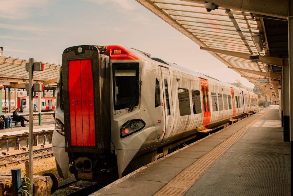 A modern commuter train is stationary at West Ham station platform, with its sleek, silver exterior featuring red accents along the carriages. The platform is partially covered by a roof with a metal 